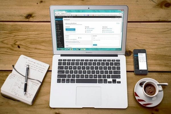 Closeup overhead of wooden desk and Laptop showing WordPress next to phone and coffee cup and notbook and pen